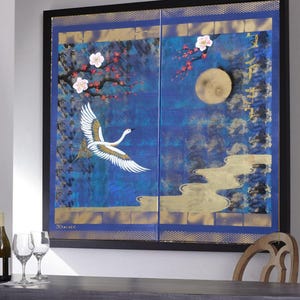 a large, colorful painting of a crane in flight against a night sky backdrop, with cherry blossoms and a full moon. The painting is framed and hangs on a wall, with a wooden chair and a bottle of wine visible in the foreground.