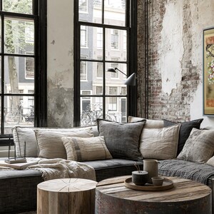 a cozy living room with a large window overlooking a city street, featuring a brick wall and a painting of a cherry blossom tree. The room is furnished with a gray sectional sofa, a wooden coffee table, and a wooden stool. A framed picture of a cherry blossom tree hangs on the wall, adding a touch of nature to the urban setting.