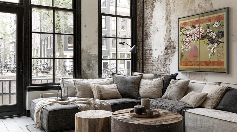 a cozy living room with a large window overlooking a city street, featuring a brick wall and a painting of a cherry blossom tree. The room is furnished with a gray sectional sofa, a wooden coffee table, and a wooden stool. A framed picture of a cherry blossom tree hangs on the wall, adding a touch of nature to the urban setting.