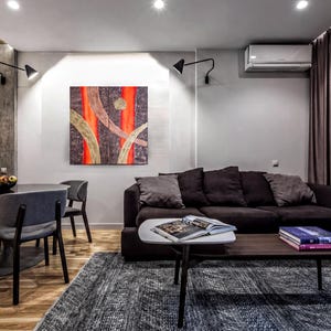 a modern living room with a black sofa, a wooden coffee table, and a dining table with chairs. The room has a neutral color scheme with gray walls and a gray rug. There is a large abstract painting on the wall, and a window with curtains is visible. The room also includes a kitchen area with a refrigerator and a sink.
