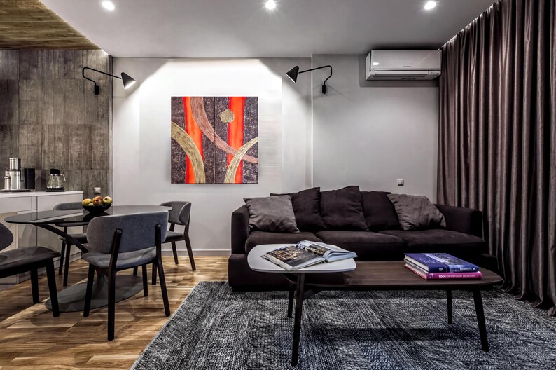 a modern living room with a black sofa, a wooden coffee table, and a dining table with chairs. The room has a neutral color scheme with gray walls and a gray rug. There is a large abstract painting on the wall, and a window with curtains is visible. The room also includes a kitchen area with a refrigerator and a sink.