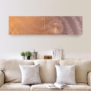 a cozy living room with a beige sofa adorned with two gray throw pillows. Above the sofa, there is a long rectangular wall art piece depicting a spiral pattern in shades of orange and brown. The wall art is positioned above a shelf that holds several books and a small plant.