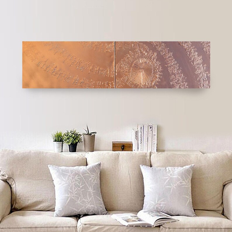 a cozy living room with a beige sofa adorned with two gray throw pillows. Above the sofa, there is a long rectangular wall art piece depicting a spiral pattern in shades of orange and brown. The wall art is positioned above a shelf that holds several books and a small plant.
