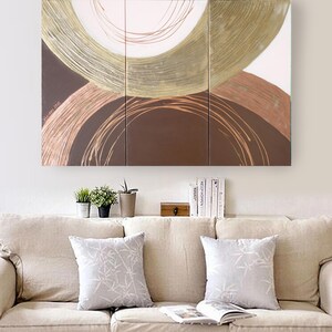 a cozy living room with a beige sofa adorned with two gray throw pillows. Above the sofa, there is a large abstract painting with a circular design in shades of brown and gold. On the wall below the sofa, there is a shelf holding several books and a small wooden box.