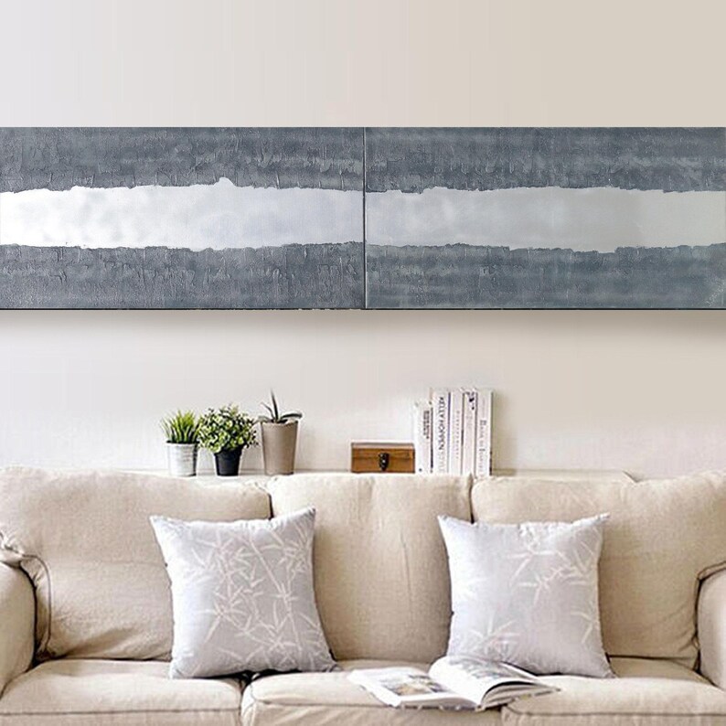 a cozy living room with a beige sofa adorned with two patterned throw pillows. Above the sofa, there is a large abstract painting with a gray and white color scheme. On the wall, there is a shelf holding several books and a small wooden box.