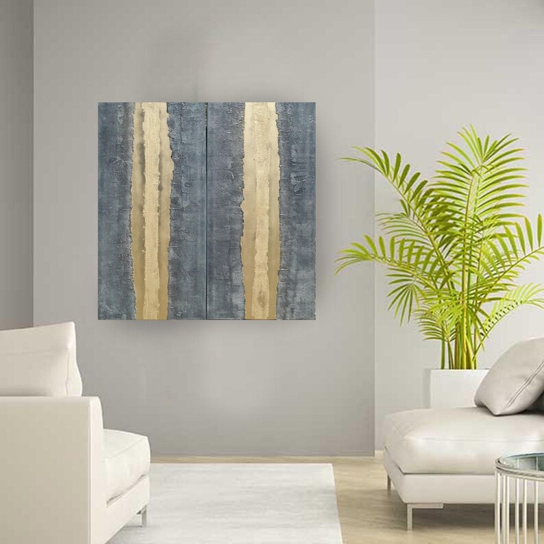 a modern living room with a large abstract painting featuring vertical lines in shades of blue and gold hanging on the wall. The room is furnished with a white sofa and a potted plant, creating a serene and stylish atmosphere.