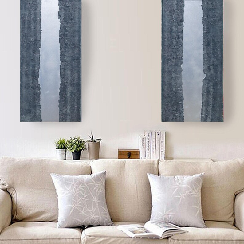 a cozy living room with a beige sofa adorned with two gray throw pillows. Above the sofa, two large paintings hang on the wall, one depicting a vertical stripe and the other a horizontal stripe. On the wall, there is a small wooden shelf holding a few books and a plant.