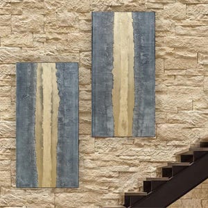 two abstract paintings hanging on a brick wall, with a staircase visible in the background.
