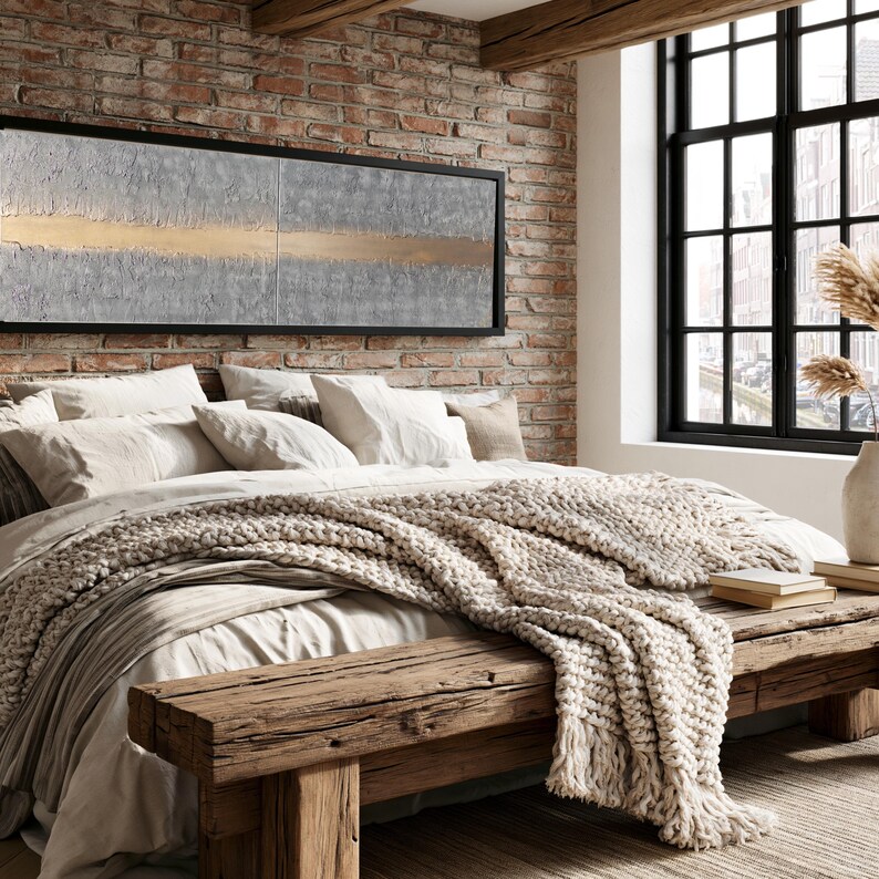 a cozy bedroom with a large bed, a wooden bench, and a brick wall with a large window. The bed is covered in white bedding and has a large blanket draped over it. The wooden bench is positioned next to the bed, and a vase is visible on the windowsill. The brick wall adds a rustic touch to the room, and the large window allows natural light to fill the space.