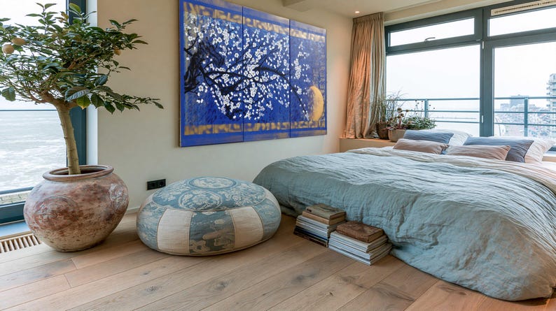 a serene bedroom with a large window overlooking the ocean, a blue and white patterned ottoman, a large painting on the wall, and a stack of books on the floor.