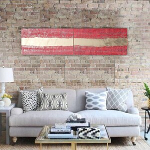 a modern living room with a gray sofa adorned with patterned throw pillows, a glass coffee table, and a brick wall backdrop. Above the sofa, there is a large, colorful abstract painting in red and white.