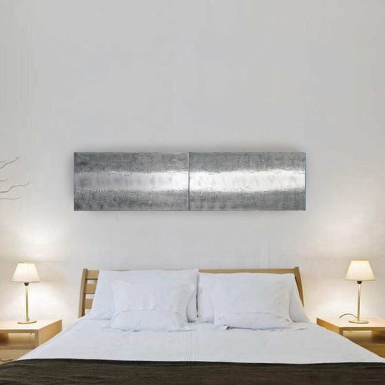 a modern bedroom with a large bed, white bedding, and two nightstands with lamps on either side. Above the bed is a large, metallic, rectangular artwork or painting.