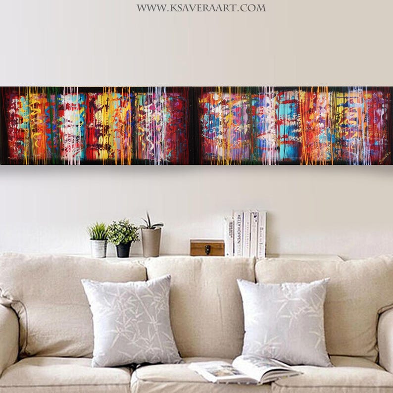 a long, colorful abstract painting hanging on a wall above a beige couch with two white throw pillows. The painting has a vibrant and dynamic design with bold lines and a mix of warm and cool colors.