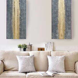 a cozy living room with a beige sofa adorned with two white throw pillows. Above the sofa, there are two large abstract paintings with vertical lines in shades of blue and gray, creating a striking visual contrast. On the wall, there is a shelf holding several books and a small wooden box.