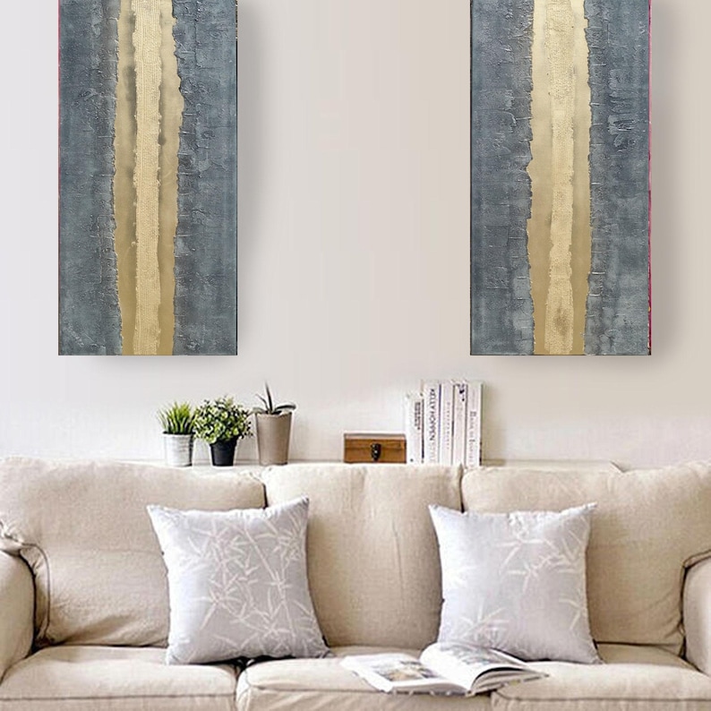a cozy living room with a beige sofa adorned with two white throw pillows. Above the sofa, there are two large abstract paintings with vertical lines in shades of blue and gray, creating a striking visual contrast. On the wall, there is a shelf holding several books and a small wooden box.