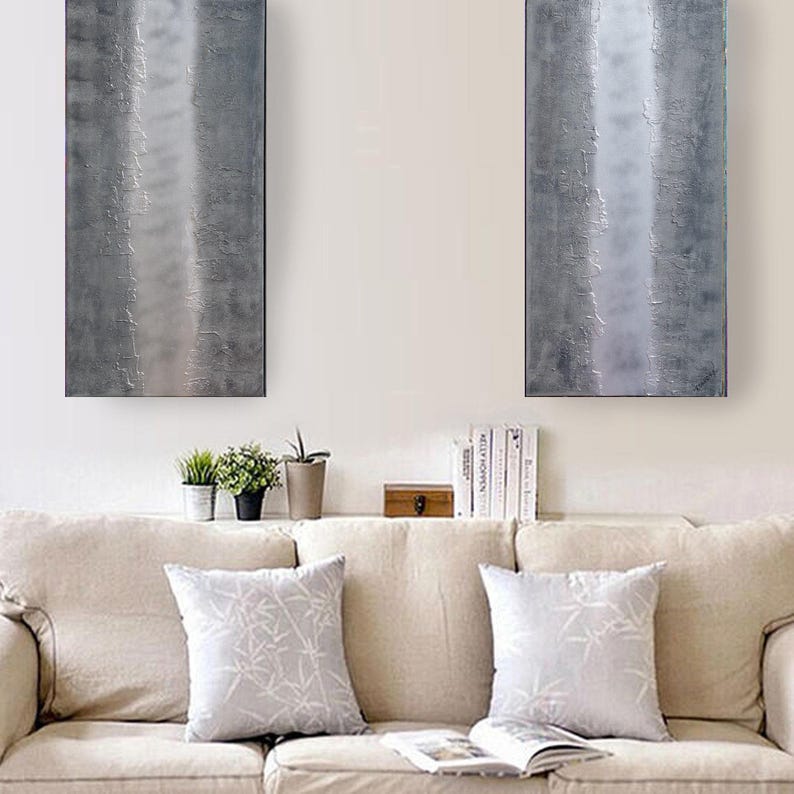 a cozy living room with a beige sofa adorned with two gray throw pillows. Above the sofa, two large abstract paintings hang on the wall, adding an artistic touch to the space. On the sofa, there are also two potted plants and a stack of books, creating a comfortable and inviting atmosphere.