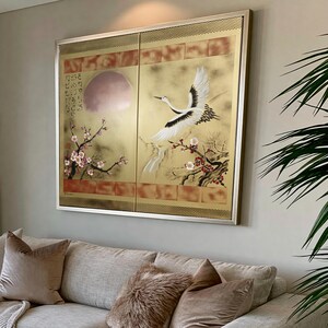 a large framed painting of a crane in flight, with a pink moon and cherry blossoms in the background, hanging on a wall above a couch with pillows.