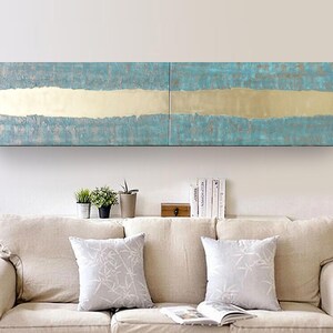 a cozy living room with a beige sofa adorned with two patterned throw pillows. Above the sofa, there is a large, abstract painting with blue and gold colors, creating a striking focal point in the room. The painting is mounted on a white wall, and the room also includes a shelf with several books and a small plant, adding to the overall aesthetic and comfort of the space.