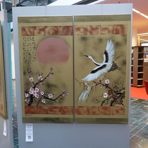 a large, colorful painting of a crane in flight, with a background of flowers and a sun. The painting is displayed on a wall, and there is a red chair visible in the background.