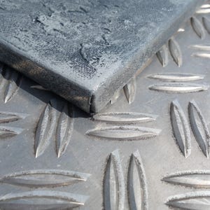 a close-up view of a metal surface with a diamond-patterned texture, featuring a dark gray or black metal piece with a rough, textured surface.