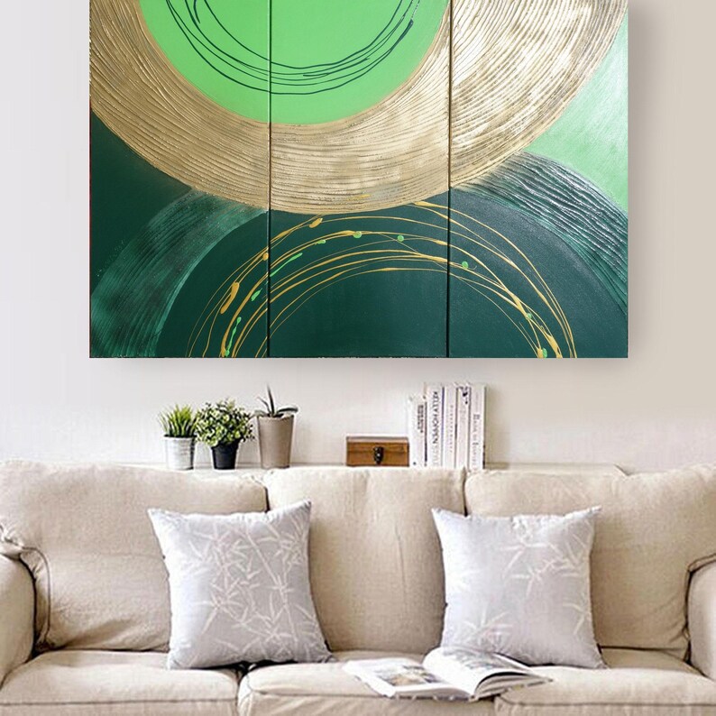 a large abstract painting hanging on a wall above a beige couch with two matching throw pillows. The painting has a green and gold color scheme with circular patterns. Below the couch, there is a shelf with several books and a small plant.