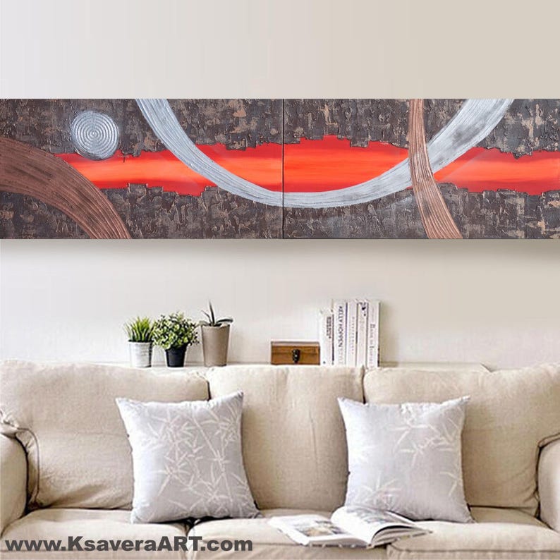 a large abstract painting hanging on a wall above a beige couch with two matching throw pillows. The painting has a dark background with bold red and white lines, creating a striking visual contrast. The couch is positioned in front of a bookshelf, which holds several books and decorative items.