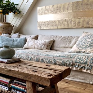 a cozy living room with a large, comfortable couch adorned with patterned throw pillows and a blue and white blanket. A wooden coffee table sits in front of the couch, holding a vase and a stack of books. The room is decorated with various plants and artwork, creating a warm and inviting atmosphere.