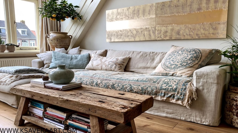 a cozy living room with a large, comfortable couch adorned with patterned throw pillows and a blue and white blanket. A wooden coffee table sits in front of the couch, holding a vase and a stack of books. The room is decorated with various plants and artwork, creating a warm and inviting atmosphere.