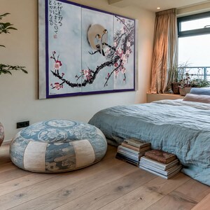a serene and cozy bedroom with a large bed, a potted plant, and a painting on the wall. The room has a large window overlooking the ocean, and the floor is made of wood. There are several books and decorative items in the room, creating a warm and inviting atmosphere.