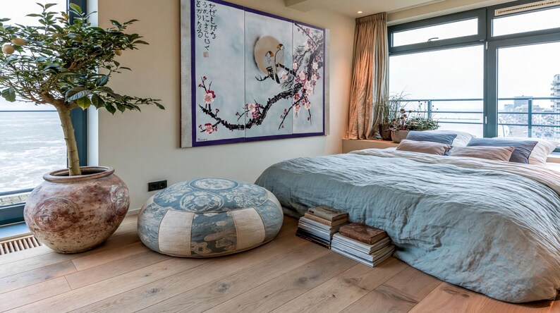 a serene and cozy bedroom with a large bed, a potted plant, and a painting on the wall. The room has a large window overlooking the ocean, and the floor is made of wood. There are several books and decorative items in the room, creating a warm and inviting atmosphere.