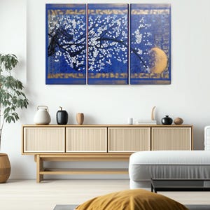 a modern living room with a large blue and gold triptych artwork hanging on the wall above a wooden cabinet. The artwork depicts a tree with white flowers against a blue background, and a yellow moon in the top right corner. The room is furnished with a white sofa, a wooden coffee table, and various decorative items such as vases and a potted plant.
