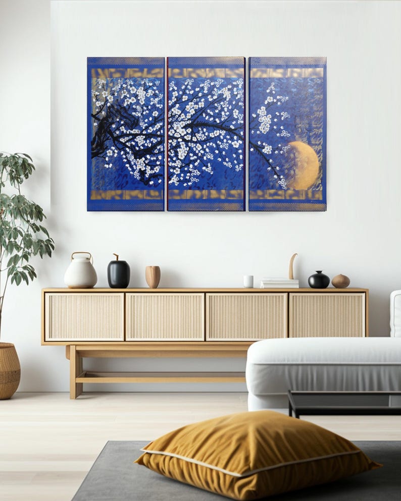 a modern living room with a large blue and gold triptych artwork hanging on the wall above a wooden cabinet. The artwork depicts a tree with white flowers against a blue background, and a yellow moon in the top right corner. The room is furnished with a white sofa, a wooden coffee table, and various decorative items such as vases and a potted plant.