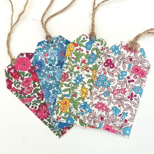 May include: Four rectangular fabric gift tags with angled top corners, each featuring a distinct floral pattern in vibrant colours including pink, blue, yellow, and green, with a natural jute twine loop attached to the top of each individual tag.