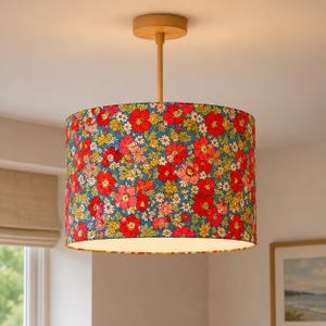 May include: A cylindrical pendant light fixture featuring a vibrant floral pattern with red, pink, and yellow blossoms set against a blue background. The drum shade is suspended from a simple, light-colored wooden ceiling mount and rod.