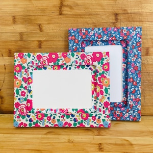 May include: Two rectangular picture frames covered in fabric with a floral pattern. One frame is oriented horizontally with a cream background, while the other is oriented vertically with a blue background, both featuring pink, orange, and green flowers.