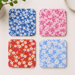May include: A set of four square coasters featuring floral patterns. One is dark blue with white flowers, one is pink with white flowers, one is orange with white flowers, and one is light blue with white flowers. Each has rounded corners.