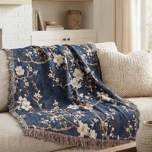 Puede incluir: Una manta azul marino con un estampado floral de flores blancas y ramas marrones. La manta está sobre un sofá beige, con un borde con flecos. Un cojín de punto color crema descansa sobre el sofá.