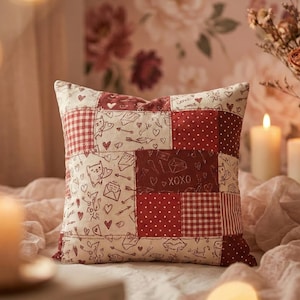 May include: A patchwork pillow with a romantic theme. The pillow features squares of red and cream fabric with patterns of hearts, cats, arrows, and the text "XOXO". The pillow is on a bed with candles and flowers in the background.
