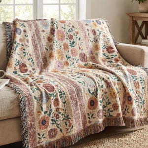 May include: A beige floral throw blanket with a fringed edge is draped over a sofa. The blanket features a repeating pattern of colorful flowers in shades of pink, yellow, and orange. The sofa is a neutral color, and a mug sits on the arm.