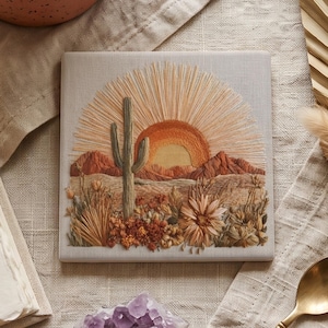 May include: A square textile art piece featuring an embroidered desert landscape. The design displays a tall saguaro cactus, a rising sun with radiating rays, and textured mountains in shades of orange, tan, and brown, accented by floral details.
