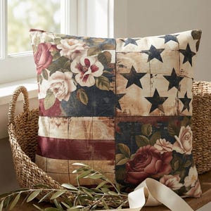 May include: Square decorative pillow with a patriotic design. The pillow features a distressed American flag pattern with floral accents, including roses and other flowers in shades of red, white, and blue. The pillow is placed on a wooden bench.