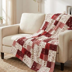 May include: A patchwork throw blanket draped over a cream-colored sofa. The blanket features a pattern of red and white squares with hearts, cats, and other small designs. The sofa sits on a woven rug in a well-lit room.