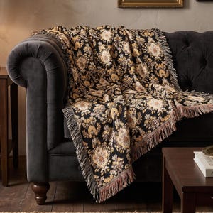 May include: A woven throw blanket featuring a black background with a repeating floral and damask pattern in shades of gold, cream, and brown. The blanket has a textured finish and is finished with decorative fringe along the edges.