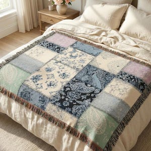 Puede incluir: Una manta de retazos con una variedad de patrones y colores, incluyendo bloques florales, paisley y lisos en tonos de azul, crema y lavanda. La manta tiene un borde con flecos y se muestra sobre una cama.