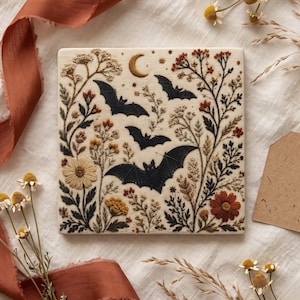 Könnte beinhalten: Eine quadratische Zierfliese mit detaillierter Stickerei-Optik, die drei schwarze Fledermäuse zeigt, welche zwischen einer Mondsichel, Wildblumen und Blattwerk in Creme, Rostorange, Senfgelb und Dunkelbraun auf hellem Grund fliegen.