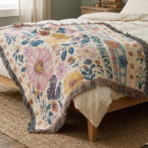 May include: A floral patterned throw blanket in cream, pink, yellow, and blue. The blanket features a large flower design with a fringed edge. The blanket is draped over a wooden bed frame and a woven rug.