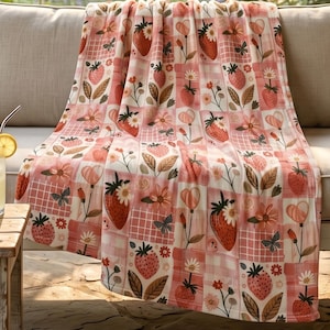 Puede incluir: Una manta estampada en rosa y blanco con un diseño de fresas. La manta tiene un diseño de patchwork con fresas, flores, mariposas y formas de corazón. La manta está sobre un sofá beige.