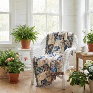 May include: A white wicker chair draped with a patchwork quilt featuring blue, beige, and red floral and plaid patterns. The quilt has a fringed edge. Potted plants and a small wooden table with a mug and book are also visible.
