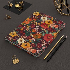 May include: A floral journal with a burgundy background and a pattern of orange, yellow, and white flowers. Gold and black pencils and gold binder clips are next to the journal.