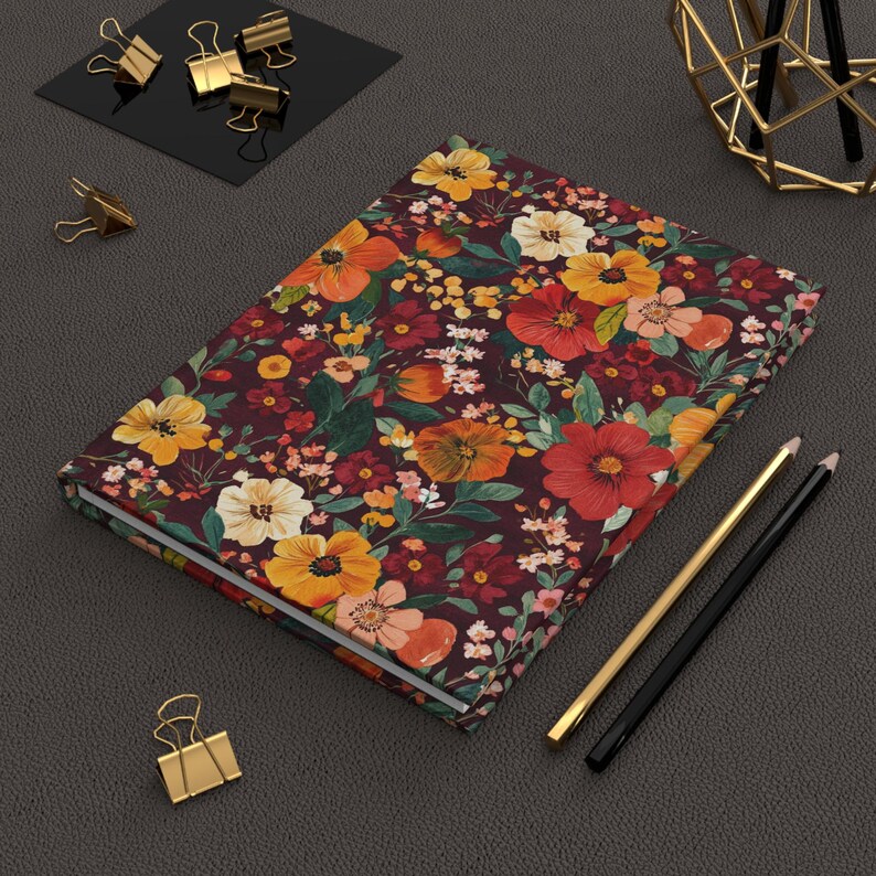 May include: A floral journal with a burgundy background and a pattern of orange, yellow, and white flowers. Gold and black pencils and gold binder clips are next to the journal.